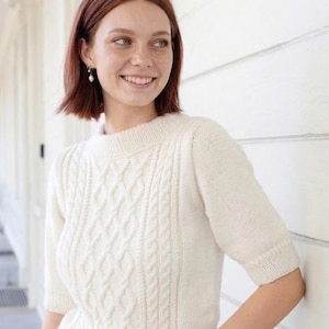 May include: Cream-colored cable knit sweater with short sleeves. The sweater features a textured cable knit design on the front and sleeves. The neckline is a crew neck. The model is wearing pearl earrings.