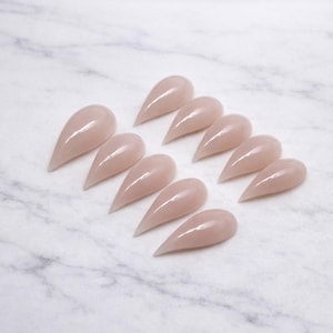 Puede incluir: Un conjunto de diez uñas artificiales en forma de almendra, de color rosa claro. Las uñas están dispuestas sobre una superficie de mármol blanco y gris. Cada uña tiene un acabado brillante y una punta afilada, ideal para manicura. Miden unos 2 cm.