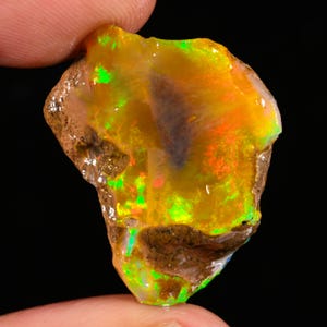 Puede incluir: Una piedra preciosa de ópalo en bruto sin pulir con una base de color amarillo dorado translúcido y destellos vibrantes de verde neón y naranja intenso. La piedra tiene una forma orgánica irregular con bordes marrones rugosos y una cara lisa.