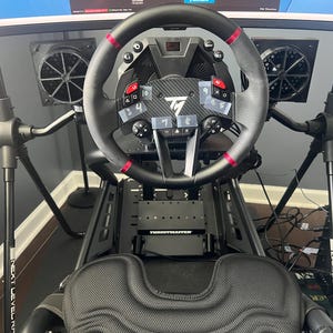 Può includere: Un cockpit per simulatore di guida nero con volante Thrustmaster dotato di accenti rossi, leve del cambio e etichette sui pulsanti. La configurazione include un sedile a guscio nero, due ventole di raffreddamento montate e un telaio in metallo.