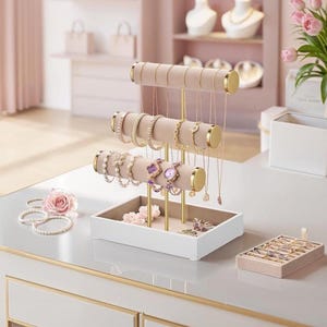 May include: A jewellery display stand with three levels for bracelets and necklaces. The stand has a white base, gold-coloured supports, and beige velvet bars. Various bracelets, necklaces, and watches are displayed on the stand.