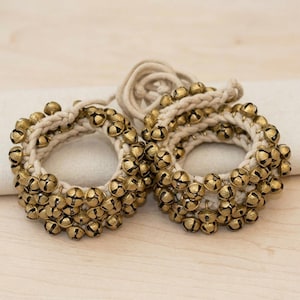 May include: Two circular anklets or wristbands constructed from thick, braided off-white cotton cord. Each band is densely covered with numerous small, spherical brass jingle bells, each featuring a cross-shaped slit on its surface.