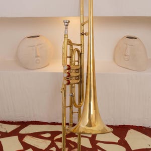 Pu&ograve; includere: Un trombone in ottone lucido con una grande campana e coulisse, posto contro un muro bianco con due vasi decorativi a forma di volto. I dettagli dello strumento sono evidenziati.