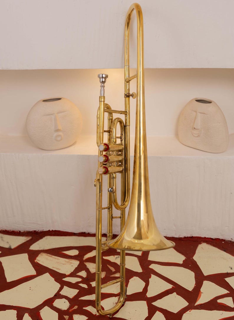 Pu&ograve; includere: Un trombone in ottone lucido con una grande campana e coulisse, posto contro un muro bianco con due vasi decorativi a forma di volto. I dettagli dello strumento sono evidenziati.