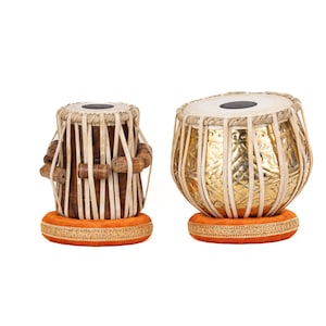 Op de afbeelding: Twee tabla-drums, traditionele Indiase percussie-instrumenten, worden getoond. Eén drum is gemaakt van hout met verticale touwaccenten en houten steunen. De andere drum is goudkleurig met ingewikkelde patronen. Beide drums rusten op oranje kussens.