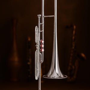 Puede incluir: Un tromb&oacute;n plateado brillante con campana acampanada y corredera. El instrumento tiene tres v&aacute;lvulas rojas y est&aacute; sobre un fondo marr&oacute;n oscuro. Un saxof&oacute;n est&aacute; borroso en el fondo.