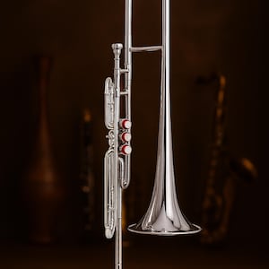 Puede incluir: Un tromb&oacute;n plateado brillante con campana acampanada y corredera. El instrumento tiene tres v&aacute;lvulas rojas y est&aacute; sobre un fondo marr&oacute;n oscuro. Un saxof&oacute;n est&aacute; borroso en el fondo.