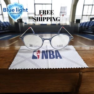 Op de afbeelding: Bril met blauwlichtbescherming met een grijs montuur en ronde glazen. De bril ligt op een witte doek met het NBA-logo. De tekst "Blue light protection" is ook aanwezig.