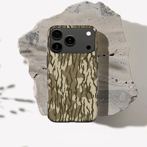 May include: A phone case with a camouflage design in green, brown, and beige tones. The case features a black camera section with three lenses and a flash. It is placed on a textured, grey stone surface.