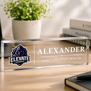Peut inclure: Une plaque de bureau rectangulaire en acrylique transparent arborant le logo Elevate Esports sur la gauche, avec le nom ALEXANDER et le titre CHIEF OPERATING OFFICER imprimés en blanc sur la droite, séparés par une ligne verticale.
