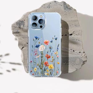 Può includere: Custodia trasparente per telefono con design floreale. La custodia ha uno sfondo trasparente e presenta fiori selvatici colorati, tra cui fiori rossi, blu, gialli e bianchi. La custodia è progettata per proteggere il telefono.