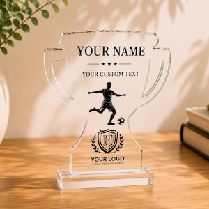 Peut inclure: Un trophée en acrylique transparent en forme de coupe, avec un texte imprimé en noir indiquant YOUR NAME, YOUR CUSTOM TEXT, YOUR LOGO et YOUR SLOGAN HERE, ainsi qu'un graphique d'un joueur de football et un emblème en forme de bouclier avec un H.