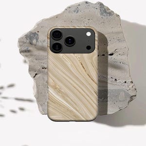 May include: A phone case with a marble design in shades of beige and gold. The case has a black camera module with three lenses and a flash. The case is resting on a rough, gray stone.