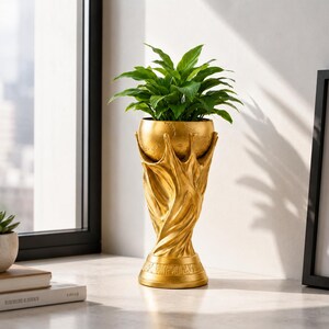 Peut inclure: Un cache-pot doré doté d'une base en forme de mains tendues ou de racines entrelacées soutenant un bol sphérique. Le bol contient une plante verte aux feuilles vives. La structure présente une finition texturée et une base circulaire large.