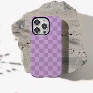 Puede incluir: Una funda de teléfono lavanda con un patrón de tablero de ajedrez. La funda tiene forma cuadrada con esquinas redondeadas y un recorte para la cámara. Descansa sobre una superficie de piedra gris rugosa.