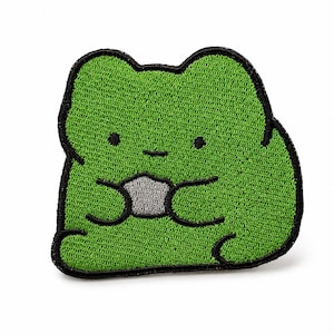 Könnte beinhalten: Ein grüner gestickter Aufnäher in Form eines sitzenden Frosches mit schwarzer Umrandung. Der Frosch hat zwei kleine schwarze Punkte als Augen, einen einfachen horizontalen Strich als Mund und hält ein kleines, hellgraues rundes Objekt in den Händen.