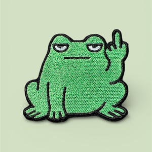 Può includere: Toppa ricamata raffigurante una rana verde con contorno nero, che mostra il dito medio. La rana ha un'espressione scontrosa. La toppa è destinata all'abbigliamento o agli accessori.