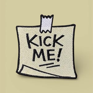 Peut inclure: Écusson brodé en forme de post-it avec l'inscription "KICK ME!" en lettres noires. Le patch est beige avec un bord noir et un détail de ruban adhésif blanc en haut.