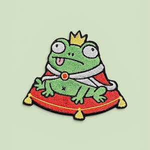 Può includere: Toppa ricamata raffigurante una rana verde con una corona d'oro e un mantello rosso con finiture bianche. La rana è seduta su un cuscino rosso con nappe dorate. La rana ha la lingua di fuori.