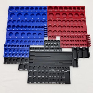 Puede incluir: Un conjunto de organizadores de herramientas modulares en azul, rojo y negro con numerosos huecos circulares para brocas o puntas. Cada sección tiene texto impreso en blanco que indica medidas imperiales y métricas específicas.