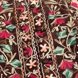 Könnte beinhalten: Ein dunkelbraunes Kleidungsstück mit einer zentralen vertikalen Knopfleiste mit passenden stoffbezogenen Knöpfen. Der Stoff ist mit aufwendigen Blumenstickereien in Magenta, Hellrosa, Creme und verschiedenen Grüntönen verziert.