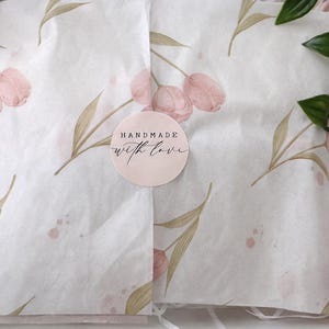 Op de afbeelding: Een roze geschenkdoos met vloeipapier met een bloemenpatroon van roze tulpen en groene bladeren. Een ronde sticker met de tekst "HANDMADE with love" staat op het vloeipapier. Groene bladeren bevinden zich in de rechterbovenhoek.