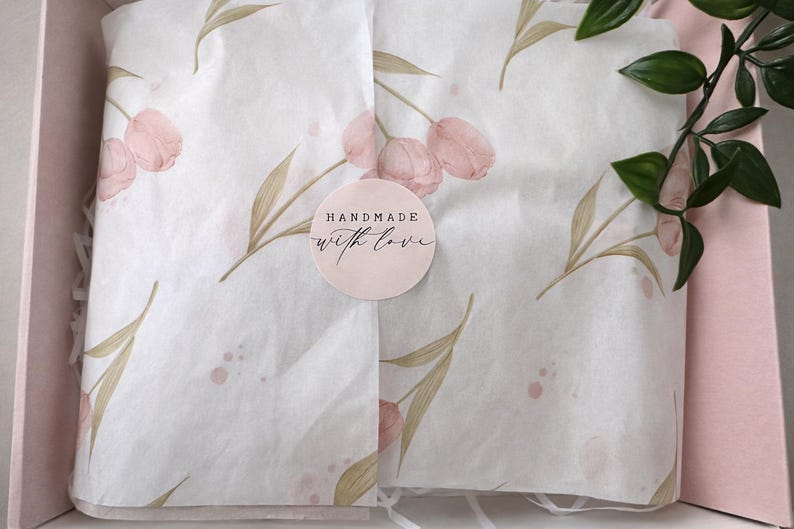 Op de afbeelding: Een roze geschenkdoos met vloeipapier met een bloemenpatroon van roze tulpen en groene bladeren. Een ronde sticker met de tekst "HANDMADE with love" staat op het vloeipapier. Groene bladeren bevinden zich in de rechterbovenhoek.