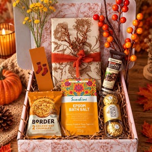 Op de afbeelding: Een geschenkdoos met een pakje Border Golden Oat Crumbles, een zakje Sunshine Epsom badzout, een tube Sadoer Coffee handcrème, twee Ferrero Rocher chocolaatjes, een bloemrijk ingepakt cadeau, een boekenlegger en decoratieve takjes.