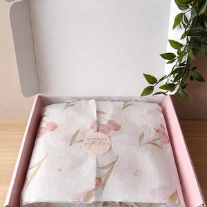 Op de afbeelding: Een roze kartonnen doos, opengeklapt om tissuepapier met een bloemenpatroon te onthullen. Het tissuepapier is om een onzichtbaar item gewikkeld. Een sticker op het tissuepapier leest "Handmade with love."