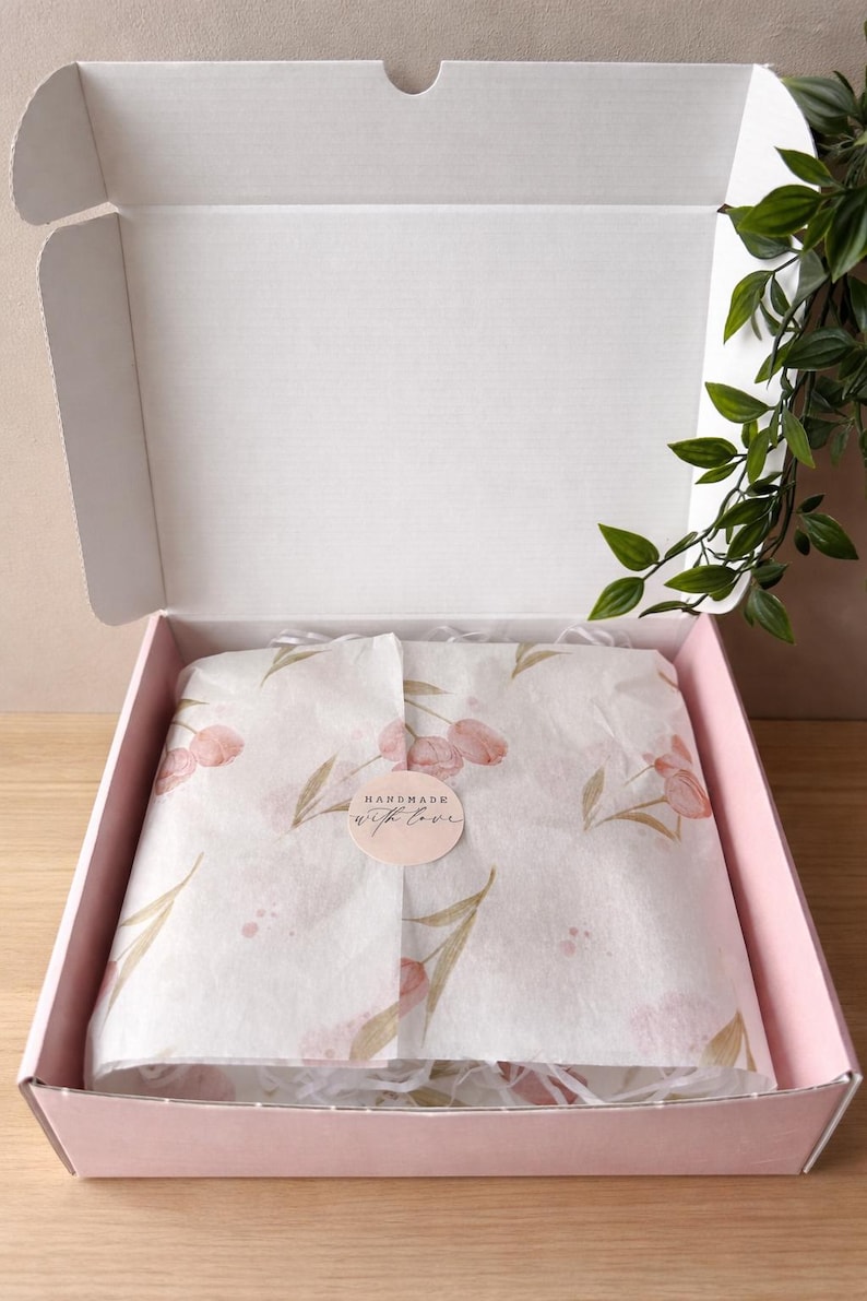 Op de afbeelding: Een roze kartonnen doos, opengeklapt om tissuepapier met een bloemenpatroon te onthullen. Het tissuepapier is om een onzichtbaar item gewikkeld. Een sticker op het tissuepapier leest "Handmade with love."
