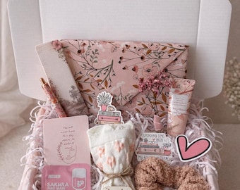 Op de afbeelding: Een roze geschenkdoos gevuld met zelfzorgartikelen. Inclusief een envelop met bloemenpatroon, sokken, een scrunchie, een gezichtsmasker, een pen en een klein boeket. De doos is gevoerd met roze papier en decoratieve vulling.