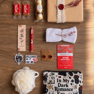 Op de afbeelding: Een platte lay-out van een cadeaupakket met diverse items op een houten oppervlak. Inclusief Biscoff-koekjes, Ferrero Rocher-chocolade, een ingepakt cadeau, een bladwijzer, een pen, stickers, oorbellen, badzout, een loofah en een make-up tas met de tekst "In My Dark Romance Era."