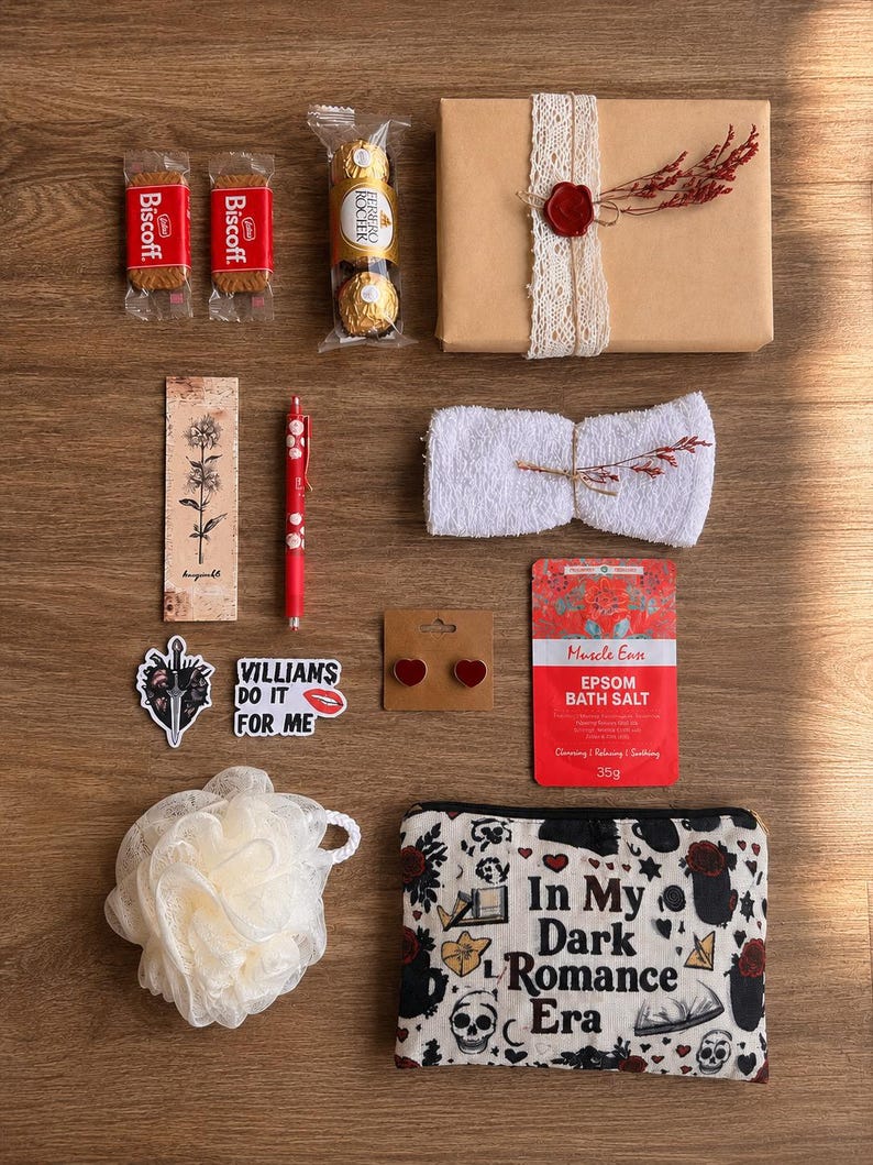 Op de afbeelding: Een platte lay-out van een cadeaupakket met diverse items op een houten oppervlak. Inclusief Biscoff-koekjes, Ferrero Rocher-chocolade, een ingepakt cadeau, een bladwijzer, een pen, stickers, oorbellen, badzout, een loofah en een make-up tas met de tekst "In My Dark Romance Era."