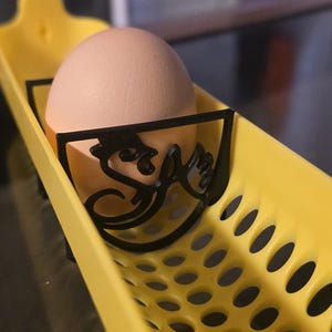 May include: A light brown egg rests in a yellow egg holder. The holder has a black decorative front with a stylized design. The holder is a tray with multiple round holes.