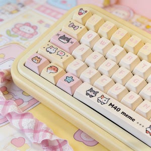 May include: A pastel yellow mechanical keyboard with custom keycaps. The keycaps feature various cat illustrations and other cute designs. The keyboard has a white space bar with the text "M40 meme".