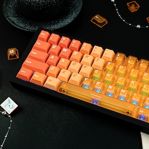May include: A mechanical keyboard with orange and transparent keycaps. The keyboard has a black frame and features standard QWERTY layout keys. The keycaps are illuminated with a warm glow, and the keyboard is angled slightly upward.