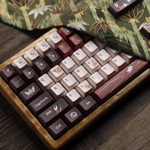 Op de afbeelding: Een set aangepaste toetsenbordtoetsen in een houten bakje. De keycaps hebben een mix van kleuren, waaronder bruin, crème en bordeaux. Sommige keycaps hebben unieke symbolen en illustraties, zoals een krab, een vogel en een gestileerd blad. De keycaps zijn gerangschikt in een standaard toetsenbordindeling.