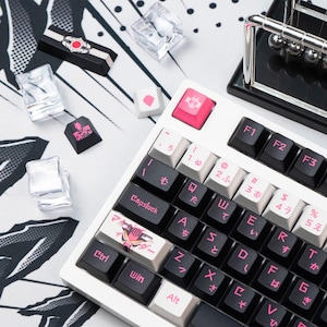 May include: A white mechanical keyboard with black and pink keycaps, some with Japanese characters. Additional keycaps and clear acrylic cubes are scattered around. A black and silver Newton's cradle is in the background.
