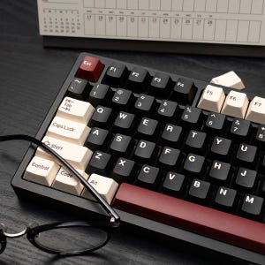 May include: Black mechanical keyboard with white and black keycaps, and a red escape key. The keyboard has a black frame and is positioned on a dark surface. A pair of black-framed glasses rests in front of the keyboard.