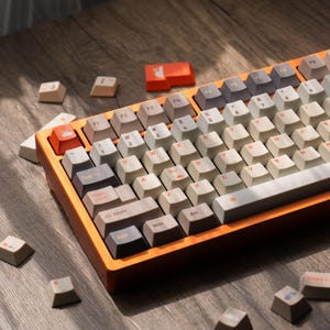Könnte beinhalten: Eine mechanische Tastatur mit einem orangefarbenen Rahmen und einer Vielzahl von Tastenkappen in neutralen Farben. Die Tasten sind in einem Standard-Layout angeordnet, wobei einige Tasten individuelle Designs aufweisen. Mehrere lose Tastenkappen liegen herum.