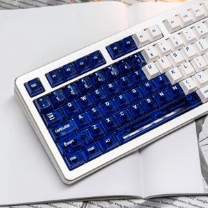 May include: A white mechanical keyboard with blue and white keycaps. The keycaps are a mix of transparent blue and white, with the letters and numbers clearly visible. The keyboard is on a white surface.