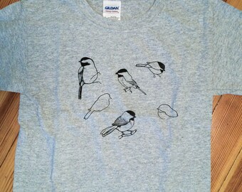May include: A gray t-shirt with a black design of multiple birds. The birds are in different positions, some are flying, some are perched, and some are looking down. The t-shirt has a Gildan tag on the front.