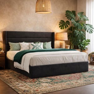 May include: A dark gray upholstered bed with a tall headboard, adorned with decorative pillows and a green throw blanket. The bed is set on a patterned rug, with a woven pendant light overhead. A large potted plant and side table complete the bedroom scene.