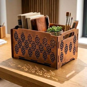 May include: A copper-toned wooden storage crate with a geometric pattern, containing books, a small plant, and art supplies. The crate, with handles, sits on a wooden desk, perfect for organizing a workspace.