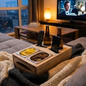 Könnte beinhalten: Ein hölzernes Sofatablett mit zwei Edelstahl-Lebensmittelbehältern, zwei Bierflaschen, einer Fernbedienung und einem Smartphone. Das Tablett befindet sich auf einem grauen Sofa mit einer Strickdecke. Die Behälter enthalten Chips und Snacks.