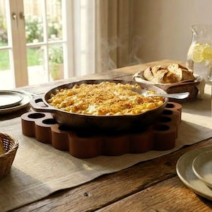 May include: A rustic wooden table setting featuring a cast iron skillet of baked macaroni and cheese on a wooden trivet. Sliced bread, plates, and a water pitcher with lemon slices complete the scene, suggesting a home-cooked meal.
