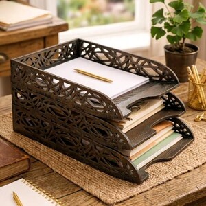May include: A three-tiered, dark brown metal desk organizer with an openwork design. The organizer holds stacks of paper and a gold pen. It sits on a woven mat on a wooden desk, suggesting an office or study setting.