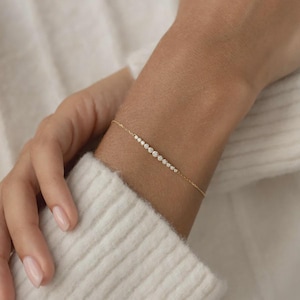 May include: A delicate gold bracelet featuring a series of small, sparkling gemstones. The bracelet is worn on a wrist, with a soft, white sweater in the background. The gemstones are evenly spaced along the bracelet's length, creating a subtle yet elegant design.