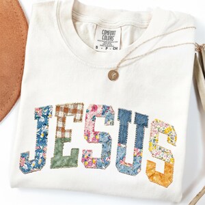 Puede incluir: Camiseta Comfort Colors color crema con la palabra "JESUS" aplicada en telas florales y estampadas de colores. Un collar de oro con un colgante descansa sobre la camisa. La camisa está doblada sobre una superficie blanca.