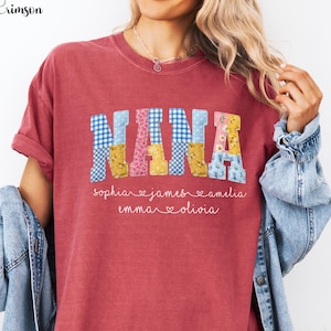 May include: A crimson red short-sleeve t-shirt featuring the word NANA in large block letters with a patchwork pattern of blue gingham, yellow floral, and pink floral prints. Below the word, five names are written in white cursive script.